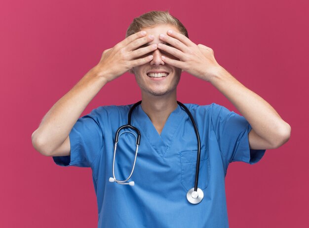 Souriant jeune médecin de sexe masculin portant l'uniforme de médecin avec le visage couvert de stéthoscope avec les mains isolés sur le mur rose