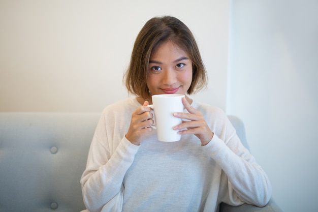 Souriant jolie femme buvant du thé et assis sur un canapé à la maison