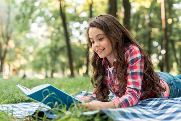 Souriant jolie fille lisant un livre en position couchée sur une couverture à carreaux au parc