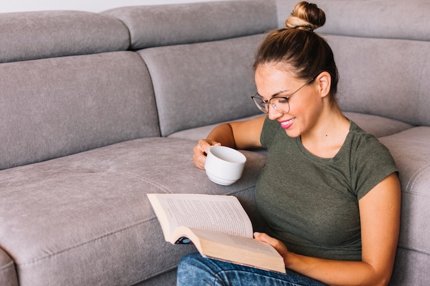 Souriant livre de lecture jeune femme tenant une tasse de café près du canapé