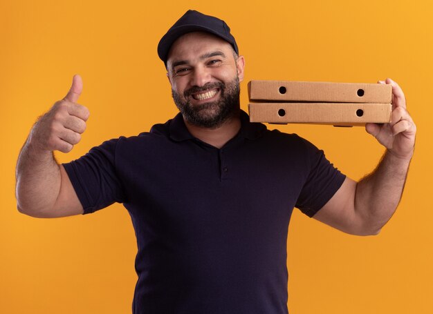 Souriant livreur d'âge moyen en uniforme et cap tenant des boîtes de pizza sur l'épaule montrant le pouce vers le haut isolé sur un mur jaune