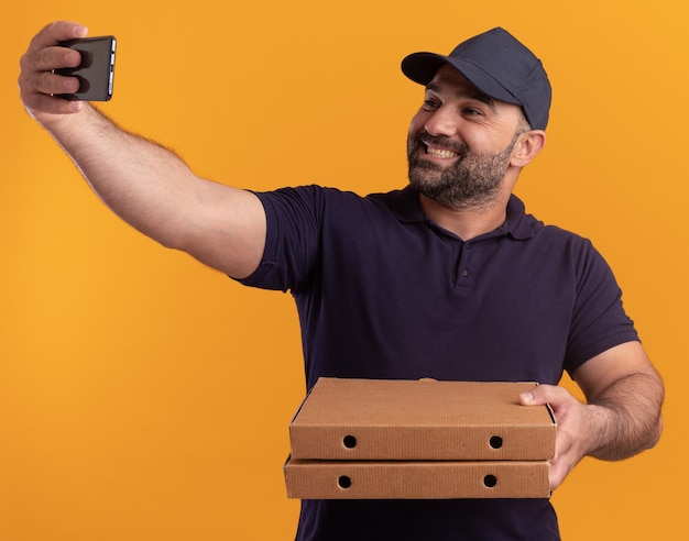 Souriant livreur d'âge moyen en uniforme et cap tenant des boîtes de pizza prendre un selfie isolé sur mur jaune