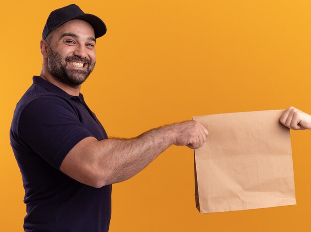 Souriant livreur d'âge moyen en uniforme et capuchon donnant un paquet de nourriture en papier au client isolé sur mur jaune