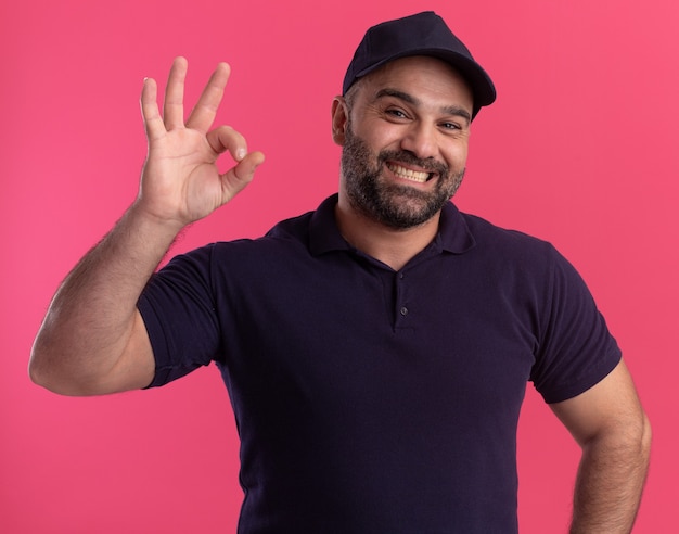 Souriant Livreur D'âge Moyen En Uniforme Et Casquette Montrant Un Geste Correct Isolé Sur Un Mur Rose