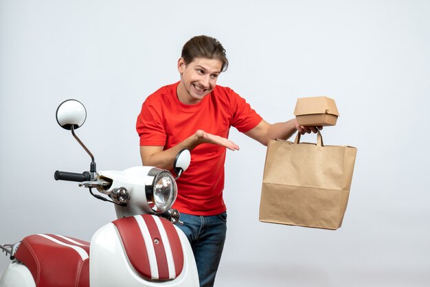 Souriant livreur en uniforme rouge debout près de scooter et montrant les commandes sur fond blanc