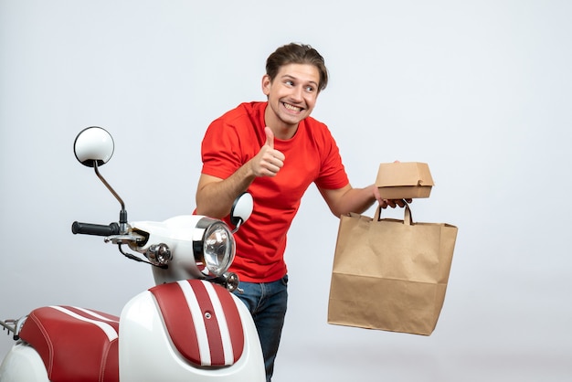 Souriant livreur en uniforme rouge debout près de scooter et tenant les commandes de cartes bancaires faisant un geste ok sur fond blanc