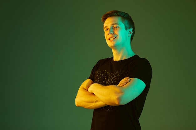 Souriant, les mains croisées. Portrait de l'homme caucasien isolé sur fond de studio vert en néon. Beau modèle masculin en chemise noire. Concept d'émotions humaines, expression faciale, ventes, publicité.