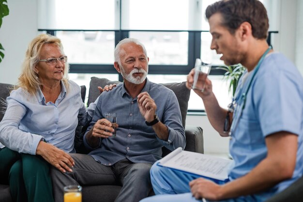 Souriant mari et femme senior parlant au médecin généraliste et lui donnant un verre d'eau lors d'une visite à domicile