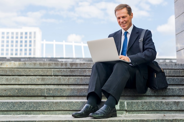 Souriant mature homme d&#39;affaires assis sur un escalier travaillant sur un ordinateur portable