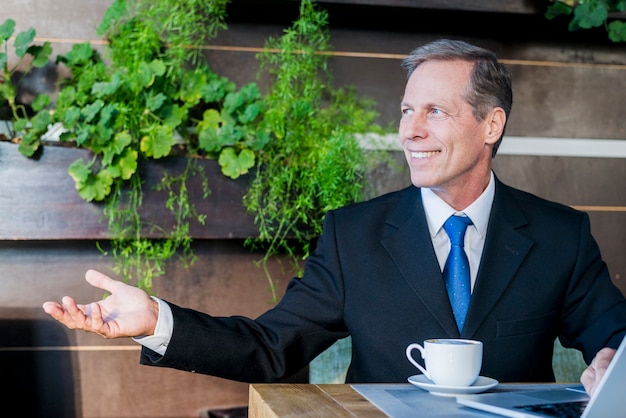 Photo gratuite souriant mature homme d'affaires fait un geste de la main avec une tasse de café et un ordinateur portable sur le bureau