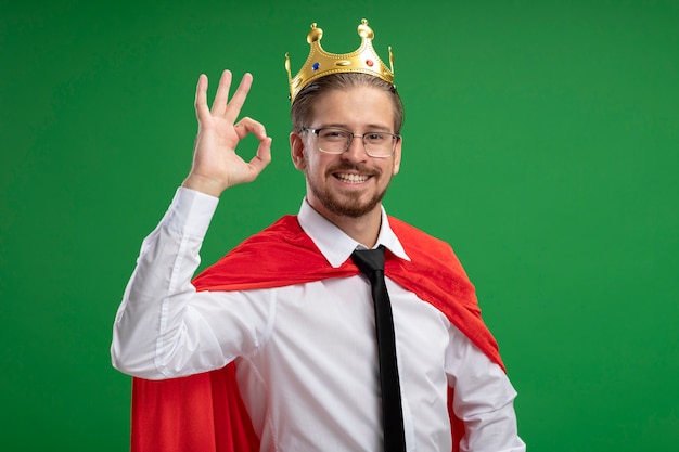 Souriant mec jeune super-héros portant couronne montrant le geste correct isolé sur vert