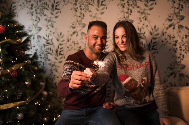 Souriant mec près dame gaie avec des lumières du Bengale près de sapin de Noël