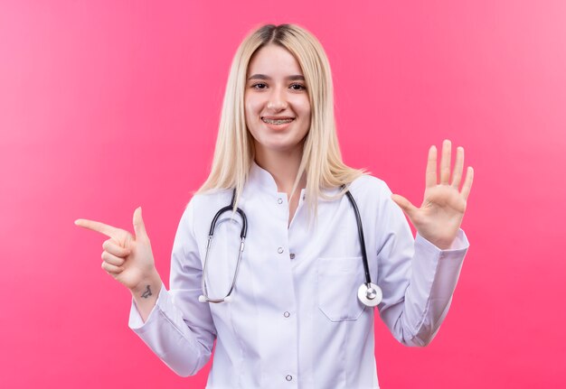 Souriant médecin jeune fille blonde portant un stéthoscope en robe médicale et orthèse dentaire montrant différents gestes sur fond rose isolé