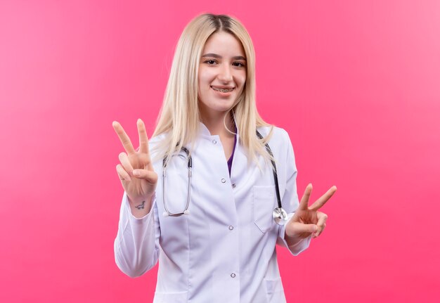 Souriant médecin jeune fille blonde portant un stéthoscope en robe médicale et orthèse dentaire montrant le geste de paix avec les deux mains sur fond rose isolé