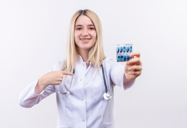 Souriant médecin jeune fille blonde portant un stéthoscope et une robe médicale en orthèse dentaire tenant des pilules se pointe sur elle-même sur fond blanc isolé