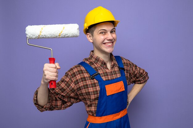 Souriant mettant la main sur la hanche jeune constructeur masculin portant un uniforme tenant une brosse à rouleau