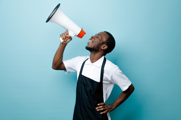 Souriant mettant la main sur les hanches jeune coiffeur afro-américain en uniforme parle sur haut-parleur isolé sur fond bleu