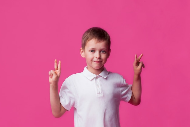 Souriant mignon petit garçon montrant le signe de la victoire sur fond rose