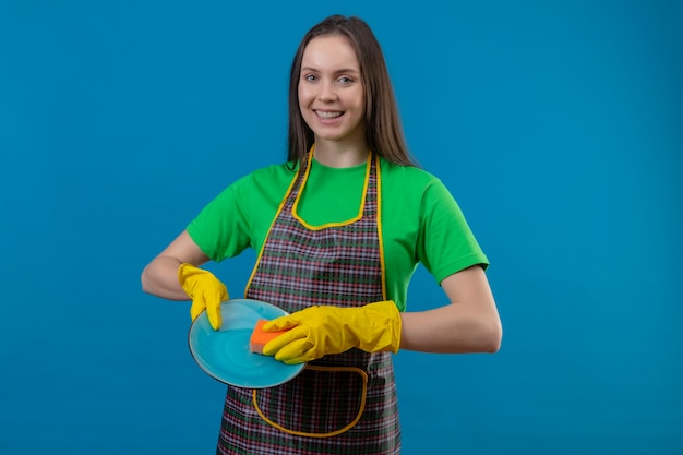 Souriant nettoyage jeune fille en uniforme dans des gants, laver la vaisselle sur fond bleu isolé