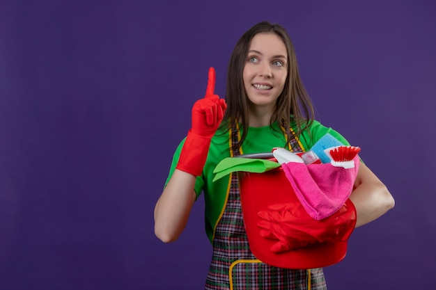 Photo gratuite souriant nettoyage jeune fille en uniforme dans des gants rouges tenant des outils de nettoyage pointe du doigt vers le haut sur fond violet isolé