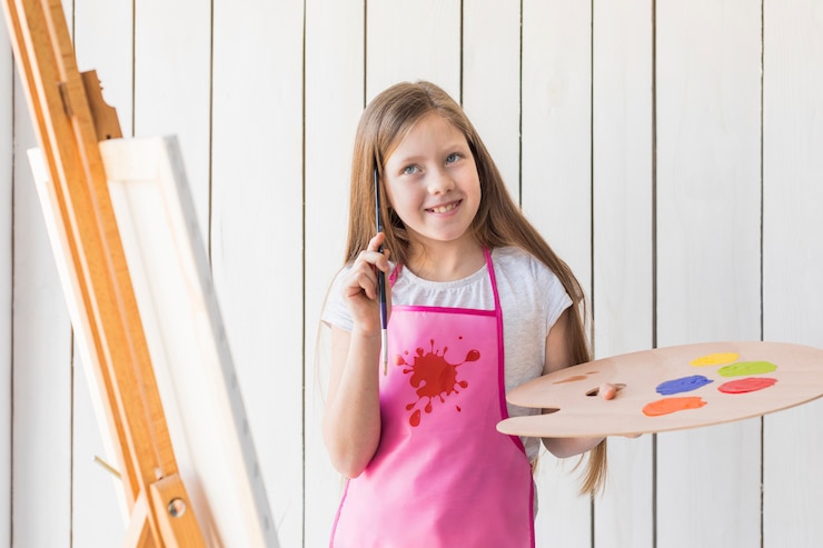 Photo gratuit souriant pensif fille tenant la palette et pinceau debout près du chevalet