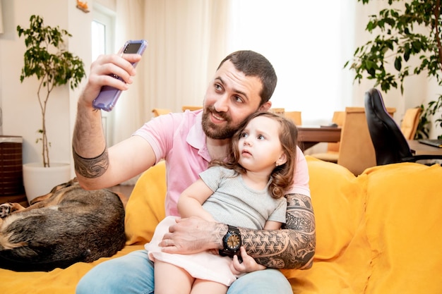 Souriant père heureux avec sa petite fille mignonne regardant l'écran du smartphone prenant la photographie de portrait selfie assis sur le canapé s'amusant ensemble concept de famille heureuse