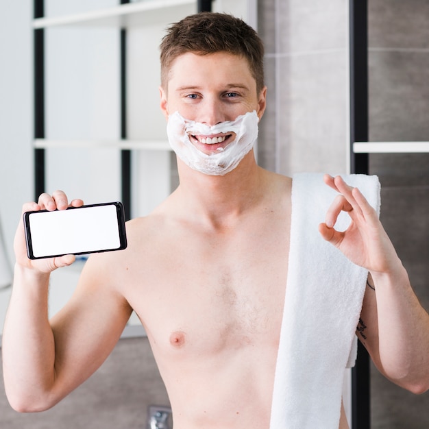 Souriant portrait d&#39;un jeune homme torse nu avec une serviette sur son épaule tenant un téléphone intelligent à la main montrant le signe ok