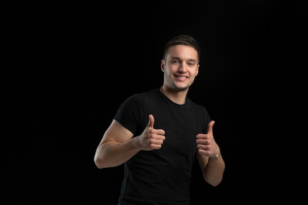 Souriant, pouces vers le haut. Portrait monochrome de jeune homme caucasien isolé sur le mur noir du studio.