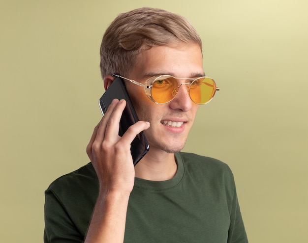 Souriant regardant côté jeune beau mec portant une chemise verte avec des lunettes parle au téléphone isolé sur mur vert olive