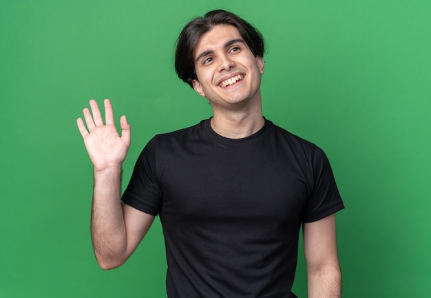 Souriant regardant le côté jeune beau mec portant un t-shirt noir montrant un geste de bonjour isolé sur un mur vert