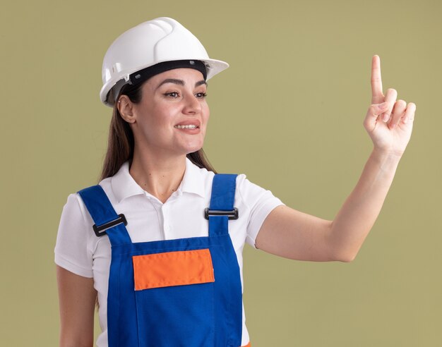 Souriant regardant côté jeune femme constructeur en uniforme montrant un isolé sur mur vert olive