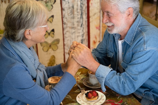 Souriant séduisant vieux couple de personnes âgées mains dans les mains regardant dans les yeux au café. cappuccino et gâteau aux fruits sur la table