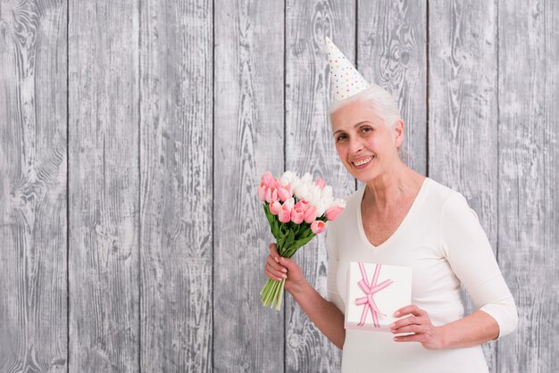 Souriant senior anniversaire femme tenant un bouquet de fleurs de tulipe et coffret cadeau en face de la toile de fond en bois