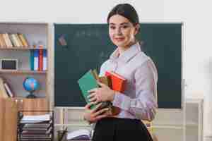 Photo gratuite souriant tenant des livres jeune enseignant debout devant le tableau noir dans la salle de classe