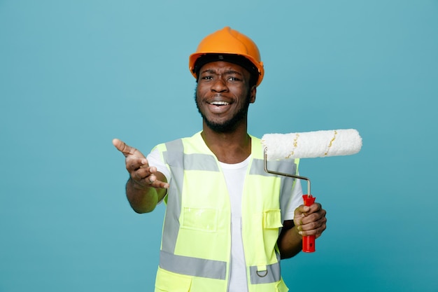 Souriant tenant la main à la caméra jeune constructeur afro-américain en uniforme tenant une brosse à rouleau isolé sur fond bleu