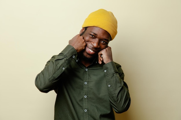 Souriant tenant les poings sur le visage jeune homme afro-américain au chapeau portant une chemise verte isolée sur fond blanc