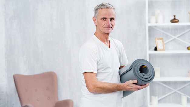 Souriant vieil homme debout à la maison avec tapis de yoga roulé