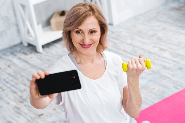 Souriant vieille femme avec des haltères prenant selfie à l&#39;aide de téléphone portable