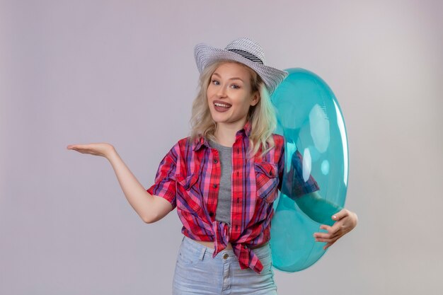 Souriant voyageur jeune fille portant une chemise rouge au chapeau tenant l'anneau gonflable pointe vers le côté sur fond blanc isolé