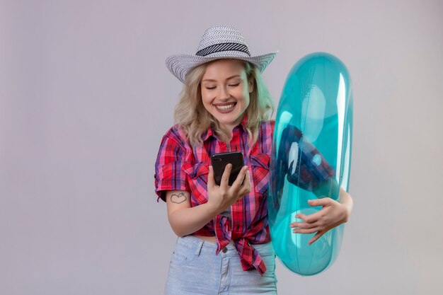 Souriant voyageur jeune fille portant une chemise rouge au chapeau tenant l'anneau gonflable à la recherche de téléphone sur fond blanc isolé