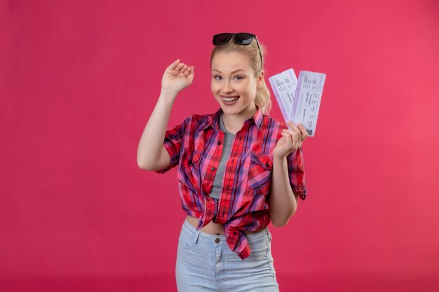 Souriant voyageur jeune fille portant une chemise rouge dans des verres tenant des billets montre la taille sur fond rose isolé