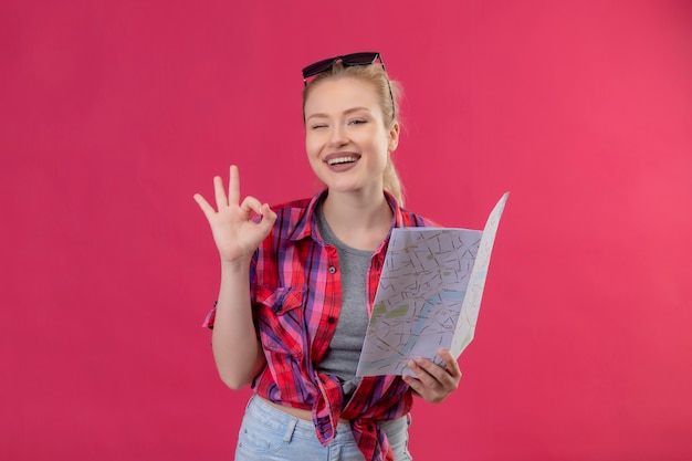 Souriant voyageur jeune fille portant une chemise rouge et des lunettes sur sa tête tenant la carte montrant le geste okey sur fond rose isolé