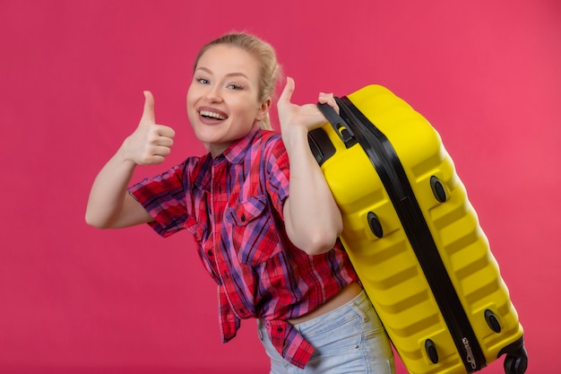 Souriant voyageur jeune fille portant une chemise rouge tenant soitcase sur le dos son pouce vers le haut sur fond rose isolé