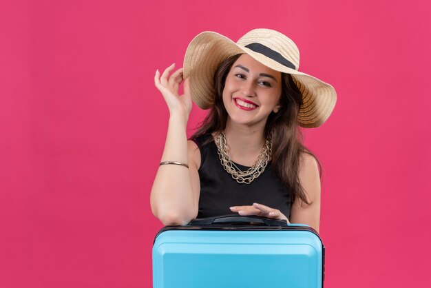 Souriant voyageur jeune fille portant un maillot de corps noir au chapeau a mis sa main sur le chapeau sur fond rouge