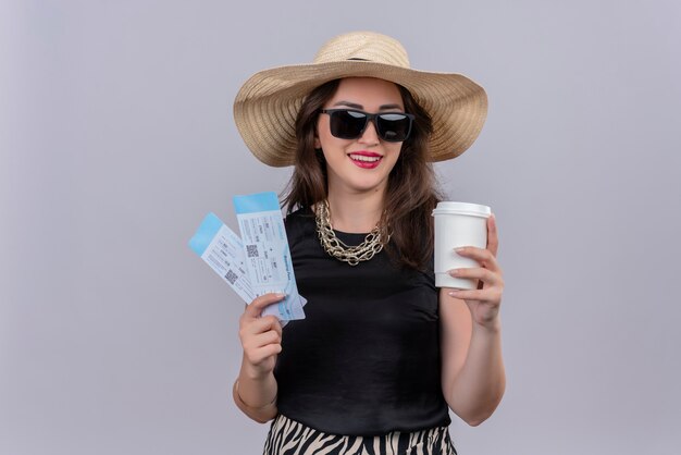 Souriant voyageur jeune fille portant un maillot de corps noir en chapeau portant des lunettes tenant des billets et une tasse de café sur fond blanc