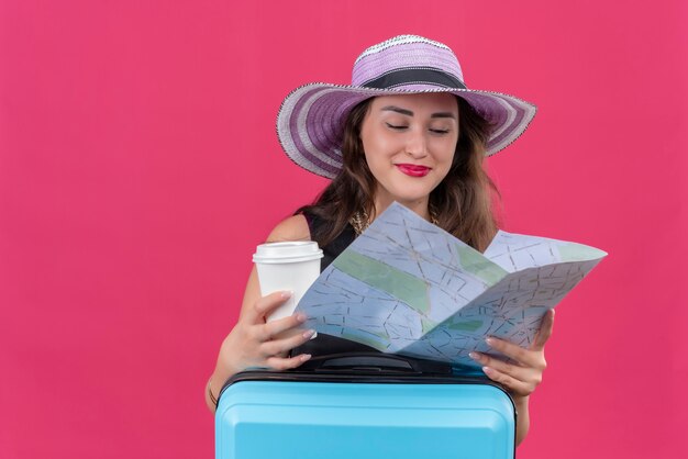 Souriant voyageur jeune fille portant un maillot de corps noir en chapeau tenant une carte et une tasse de café sur fond rouge