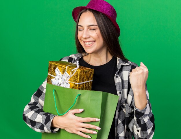 Souriant avec les yeux fermés jeune belle fille portant un chapeau de fête tenant un sac-cadeau montrant un geste oui