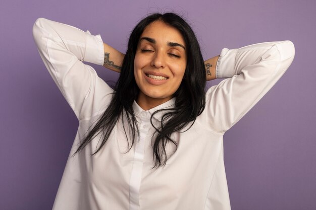 Souriant avec les yeux fermés jeune belle fille portant un t-shirt blanc tenant les mains derrière la tête isolé sur violet