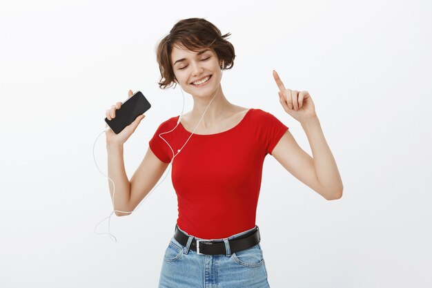 Souriante belle femme brune danse insouciante, écoute de la musique et danse dans les écouteurs, tenant le smartphone