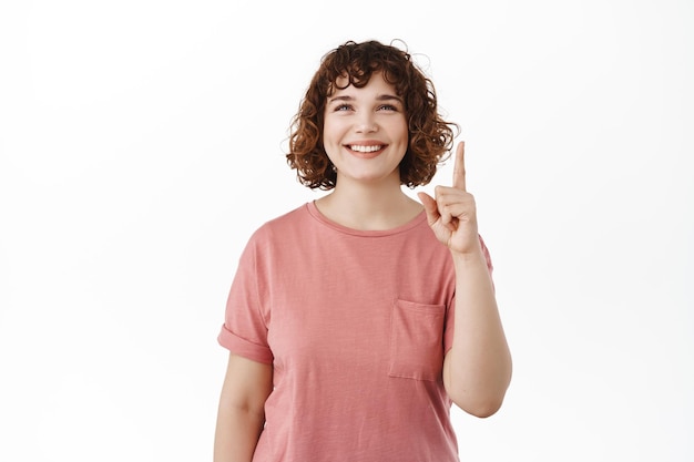 Souriante belle fille bouclée, pointant le doigt vers le haut et regardant avec un visage heureux rêveur, debout joyeuse et rêveuse sur fond blanc